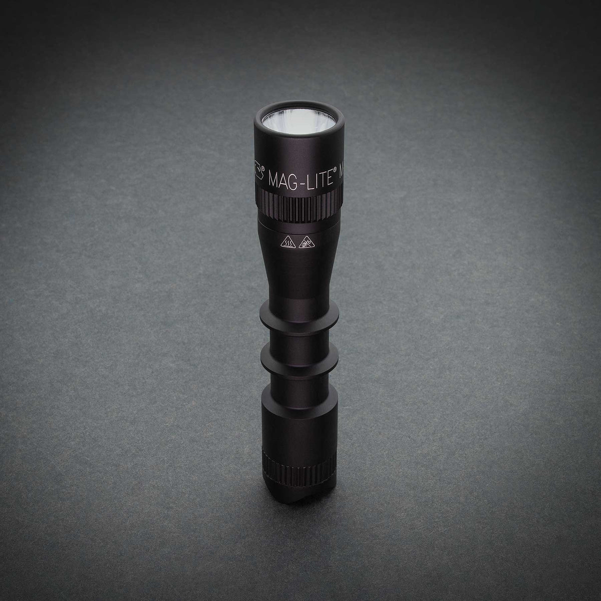 Front view of a black MAG-TAC 2 TL flashlight with plain bezel against a dark grey studio backdrop
